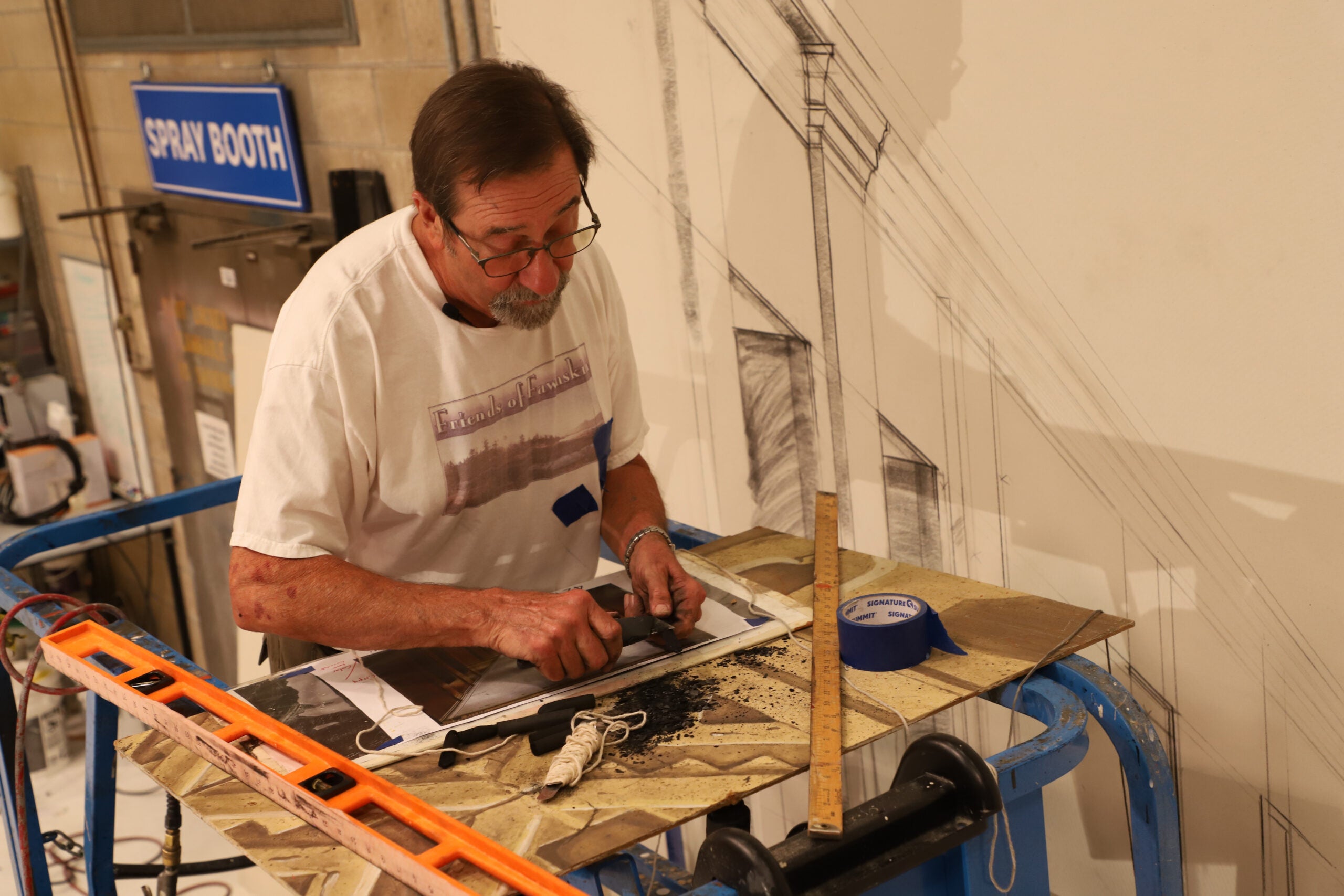 Guest scenic artist Joe Francuz working in the Hollywood Backdrops Collection studio during a visiting artist residency. The photograph documents Francuz creating scenic artwork using traditional theatrical painting techniques and drafting methods as part of educational programming for the collection.