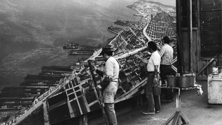 Black-and-white photograph of scenic artists Ben Resella, Fred Tuch, and Harry Tepker working on a large cityscape backdrop. The image documents collaborative scenic painting techniques used to create large-scale urban environments for film and stage productions.